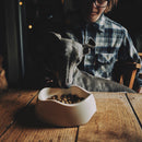 Lévrier gris mangeant des croquettes dans une gammelle en bambou blanche avec son maître en fond